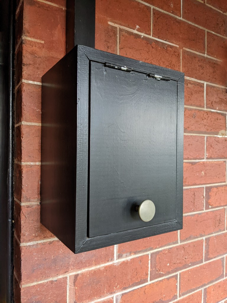 Photo of a small wooden cupboard mounted on a red brick wall. The cupboard houses the smart meter, so is about 40cm high and 30 cm wide. The cupboard is painted black, and is closed in the photo.